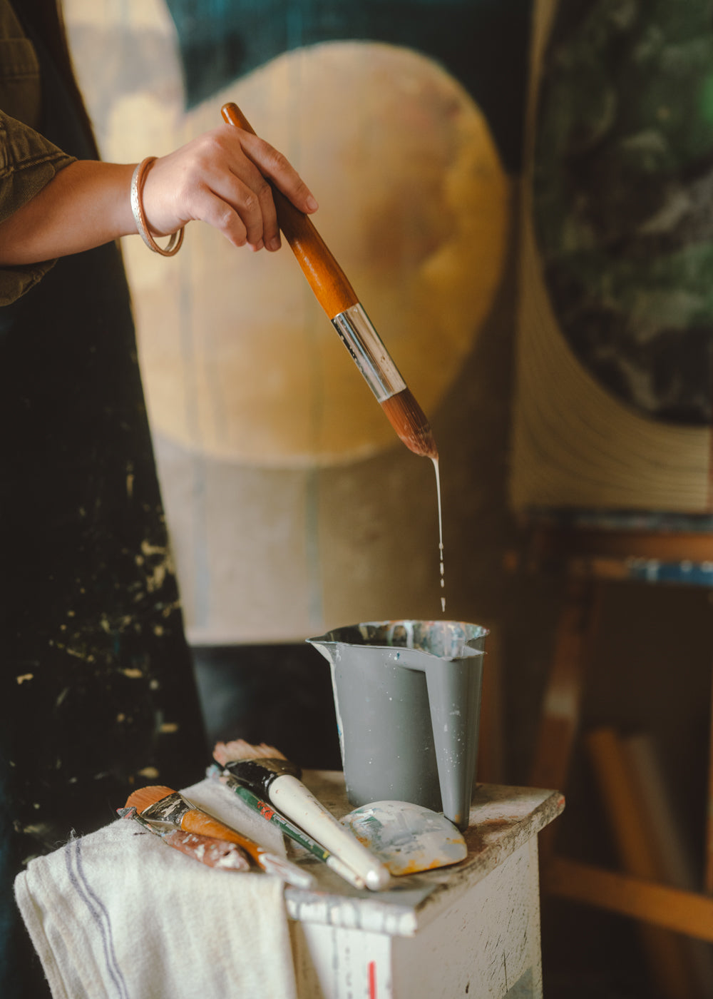 Person holding a paintbrush over a container with paint, surrounded by art supplies.