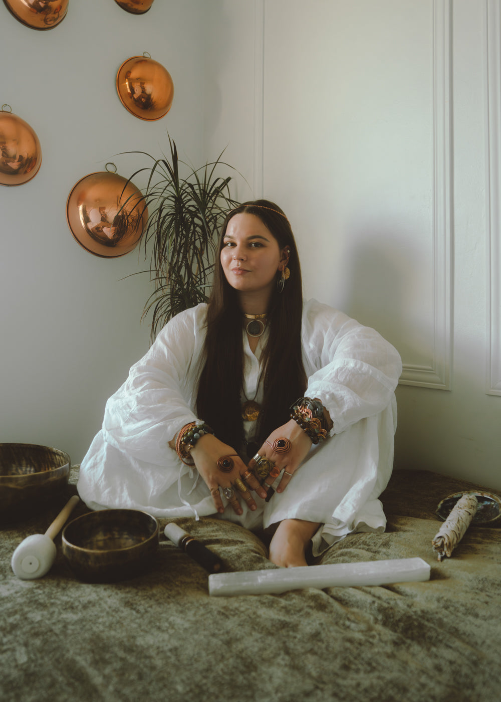 Woman in a white outfit sitting on the floor with various objects around her, including copper pots on the wall.